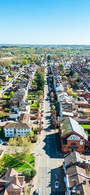 An aerial view of a suburban neighborhood featuring a long straight road lined with houses and trees, stretching into the distance under a clear blue sky.