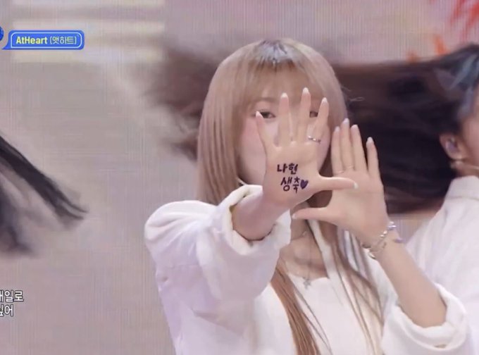 A person with long blonde hair covers their face with their hands, which have Korean text written on them, during what appears to be a live broadcast on 'AtHeart' music show.