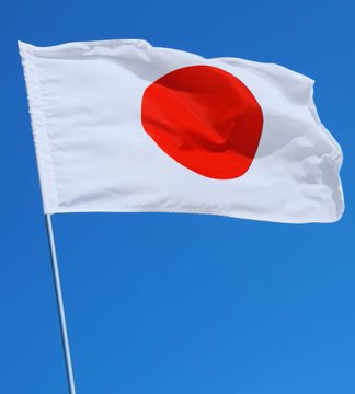 The image shows the flag of Japan—a white banner with a central red circle—waving against a clear blue sky, symbolizing the country's currency and economic context. The flag visually represents Japan as the setting for the news about Japanese firms increasingly adopting Bitcoin amid a weakening yen, with companies incorporating it into shareholder benefit programs.