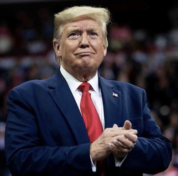 Portrait of Donald Trump in a dark blue suit with red tie, clapping his hands at what appears to be a rally.
