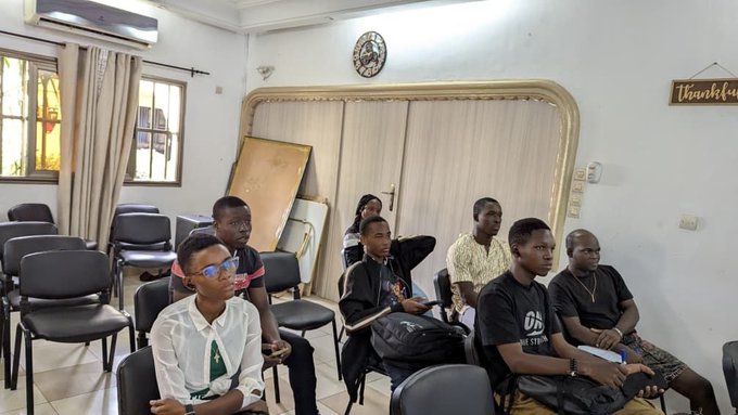 A group of young men sit attentively in black chairs inside a room with white walls and a decorative mirror; a 'Thankful' sign hangs on the wall, and a clock reads approximately 1:10.