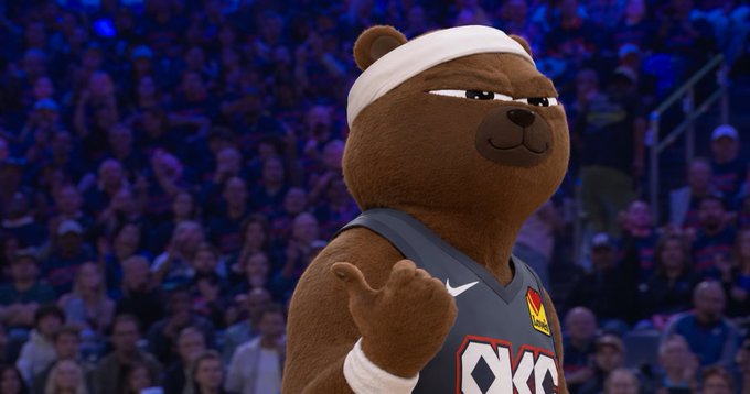 An animated brown bear character wearing a white headband, white wristband, and an OKC (Oklahoma City) basketball jersey, giving a thumbs-up while standing in front of a basketball arena crowd.