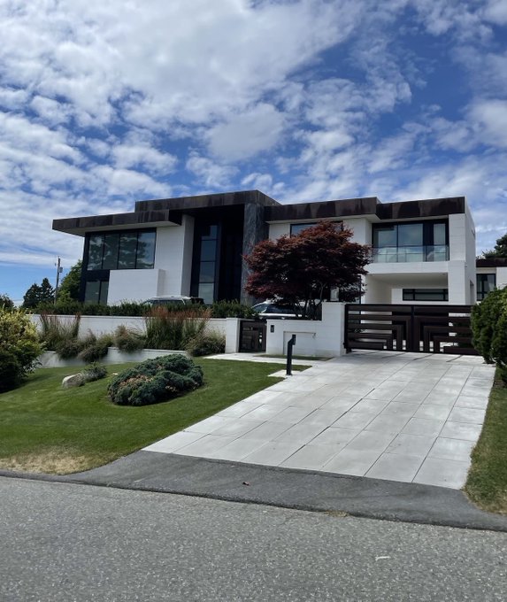 A modern, multi-level house with dark and white exterior, a tiled driveway, and a small landscaped garden under a blue sky with scattered clouds.