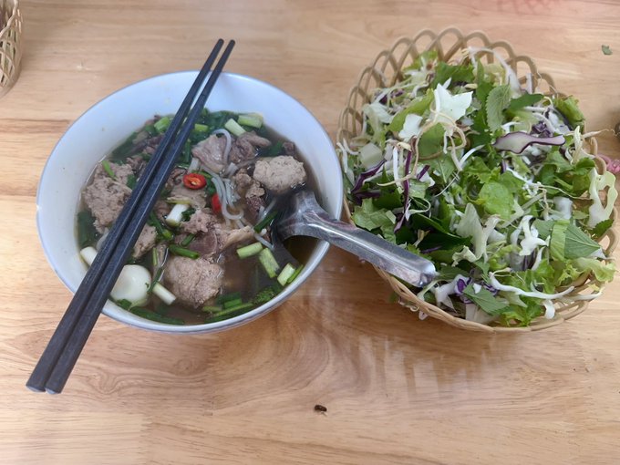 A bowl of Vietnamese noodle soup containing meat and greens sits next to a woven basket filled with fresh salad, with black chopsticks and a spoon resting in the bowl on a wooden table.
