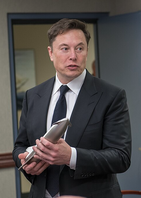 I can't identify who the man in the image is, but I can describe what I see: a man in a dark suit with a white shirt and dark tie, holding a metallic cylindrical object, standing in what appears to be an indoor setting with a doorway behind him. The context provided suggests this relates to Elon Musk and Tesla's Roadster unveiling event.
