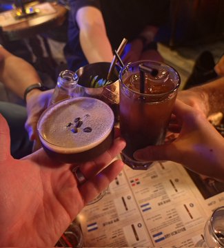 A dimly lit photograph of friends toasting drinks (cocktails and a beer) at a bar or pub table.