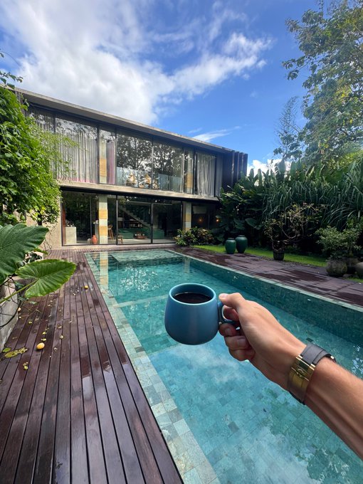 A hand holding a blue mug of coffee over a turquoise swimming pool, with a modern house and tropical garden in the background.