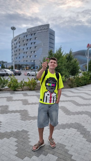 A young man in a neon yellow t-shirt and denim shorts stands on a paved plaza holding up a small object, with a building displaying 'MODULOS' in the background.