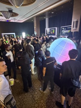 A crowded indoor event or convention with many attendees gathered around a large glowing spherical display with purple and blue colors.