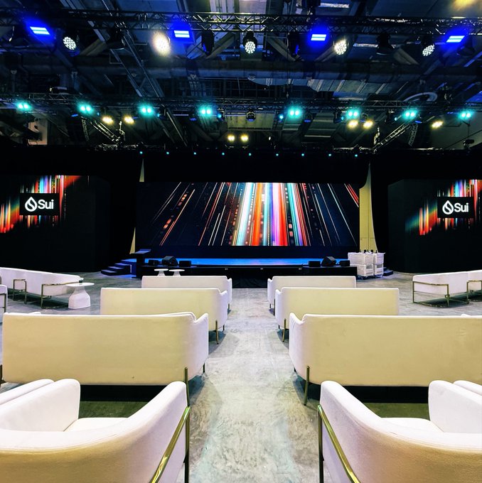 An empty auditorium or conference hall set up with cream-colored armchairs facing a stage with large screens displaying 'Sui' logos and colorful abstract designs.