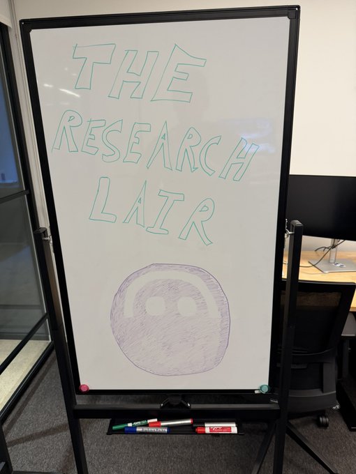 A whiteboard displays "THE RESEARCH LAIR" in teal lettering above a drawn sad face emoji, with markers resting on the tray—setting a playful scene for Bitwise Research's "coin of the week" announcement featuring AAVE in their ongoing "DeFi Decade" coverage.