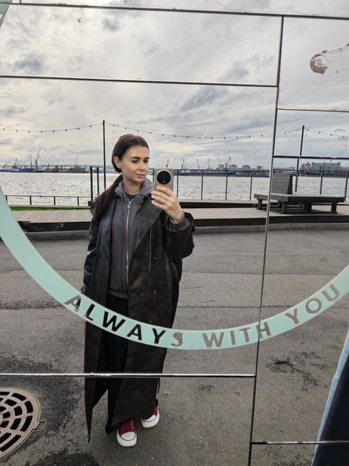 A person takes a mirror selfie with a camera, wearing a long black leather coat over a grey hoodie, reflected against a waterfront backdrop; the mirror has text reading 'ALWAYS WITH YOU'.