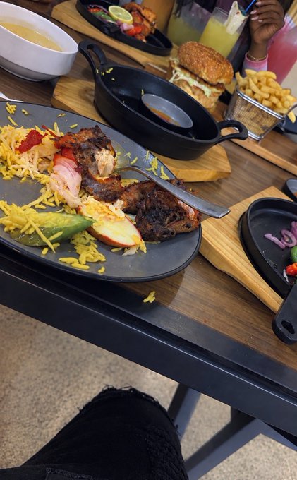 A black plate on a wooden table holds a partially eaten meal of grilled meat, yellow rice, and vegetables, with a cast iron skillet, a burger, fries, and drinks nearby.