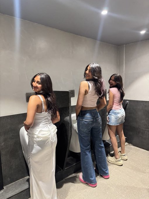 Three young women stand side by side at urinals in a public restroom, with the woman on the left smiling back at the camera in a white dress, the middle one in jeans laughing, and the right one in a pink top and denim shorts looking on. The image humorously references a "shart" (meme slang for an unexpected bowel accident) when nervous, tying into the post's anxious anticipation of a "$BASE TGE" (token generation event) announcement tomorrow.