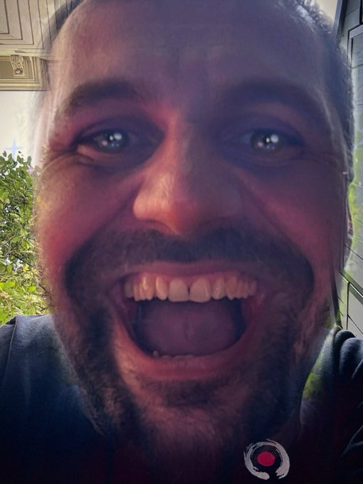 A close-up selfie of a smiling bearded man with a wide open mouth, with a small red dot logo visible in the bottom right corner.