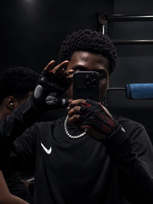 A young man takes a mirror selfie while wearing black and red Adidas gloves and a black Nike t-shirt.
