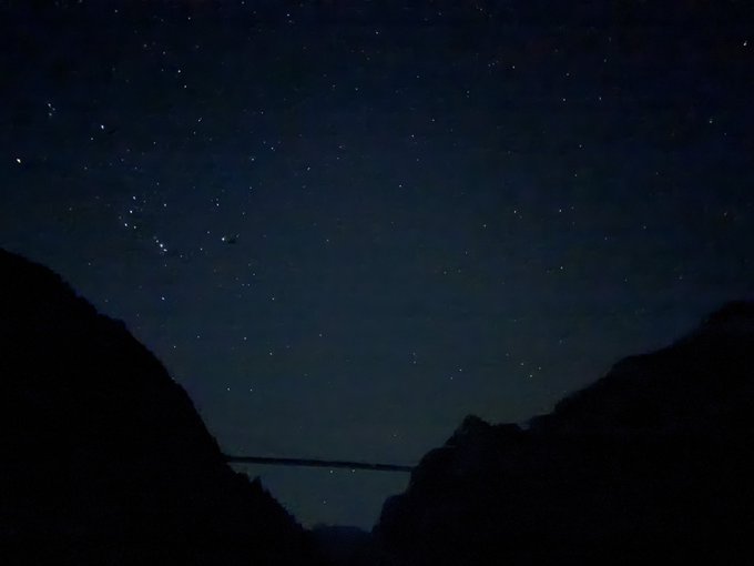 A nighttime photograph of a starry sky with silhouettes of dark mountains or hills in the foreground, possibly featuring a bridge or dam structure between them.