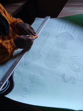 A person in an orange jacket uses a yellow crayon to draw on a large light blue poster featuring sketched letters and the words "THANK" and "SNOI".