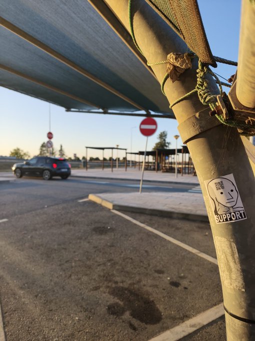 A close-up photo of a sticker on a metal pole reading 'SUPPORT' with a line-drawn face; in the background is a parking lot with a 'no entry' sign and a passing car.