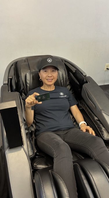 A smiling woman wearing a dark cap and 'CRYPTO WOLF' t-shirt, sitting in a black massage chair and holding up a small green card.