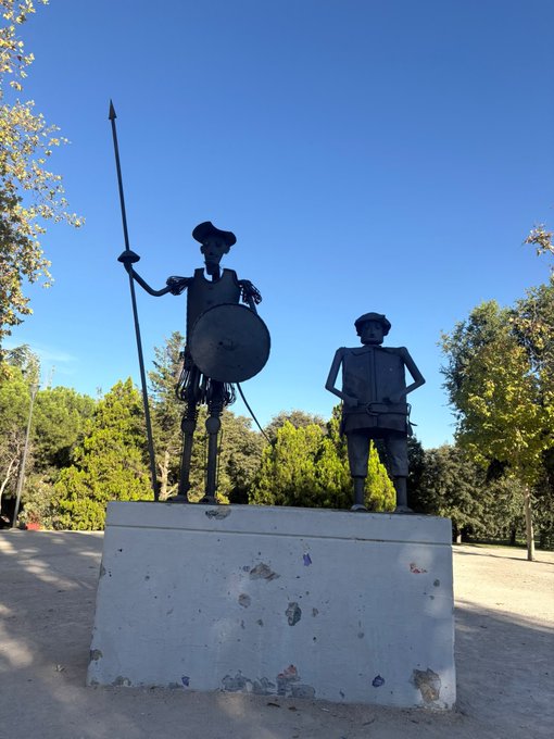 A metal sculpture of Don Quixote and Sancho Panza mounted on a white concrete pedestal, set outdoors under a clear blue sky with trees in the background.