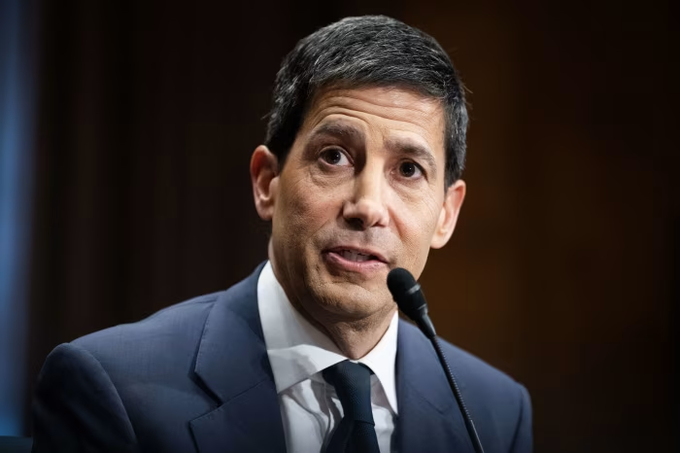 A man with dark hair, wearing a navy blue suit, white shirt, and dark tie, speaks into a microphone during what appears to be a formal hearing or testimony. The image corresponds to the post's reference to Federal Reserve Chair Kevin Warsh preparing to deliver his Monetary Policy Report testimony.