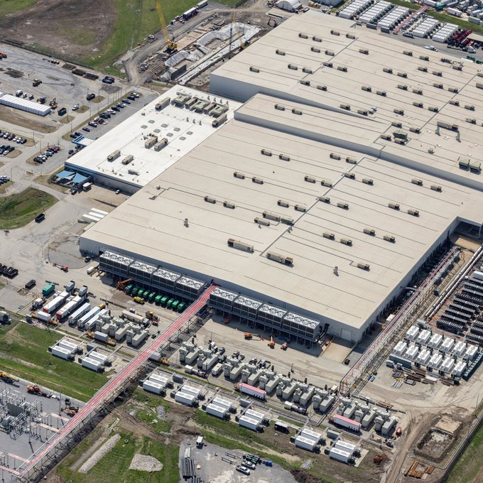 An aerial view shows a large industrial facility with expansive rooftops, surrounded by storage tanks and construction materials.