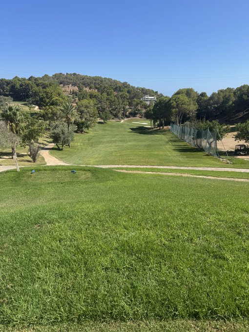 A pristine golf course stretches out under a clear blue sky, with lush green fairways framed by trees and a netting fence on the right side. The serene landscape pairs perfectly with a casual "Good afternoon" greeting, capturing a peaceful moment on the course.