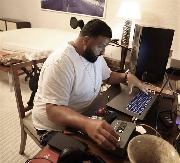 A bearded man wearing a white t-shirt sits at a desk working on a laptop, with studio monitors, a microphone, and other equipment around him.