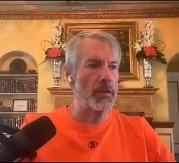 A bearded man in an orange shirt with a Bitcoin logo speaks into a microphone during what appears to be a video interview, with an ornately decorated room featuring framed artwork and floral arrangements visible in the background. The image relates to the post as it shows Michael Saylor, the MicroStrategy founder known for his Bitcoin advocacy, likely making the quoted statement about holding onto Bitcoin versus his company's strategy.