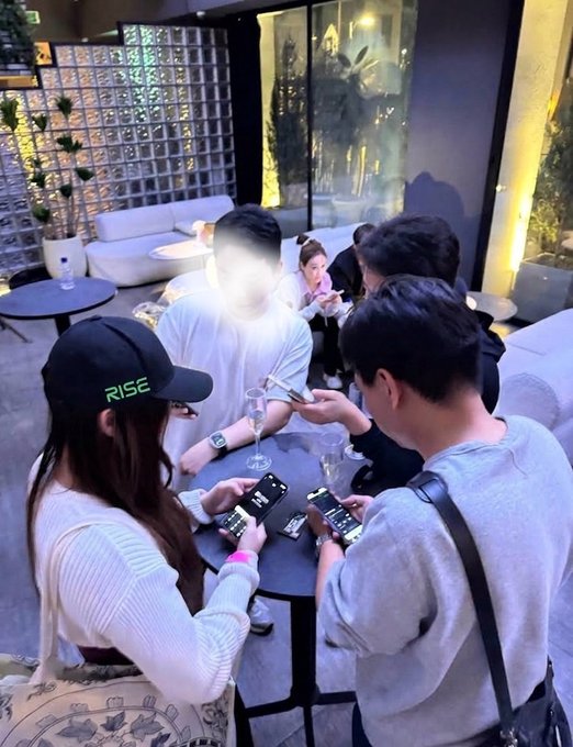 A group of people sitting around a small table at an outdoor cafe, focused on their smartphones, with one person wearing a black "RISE" cap.