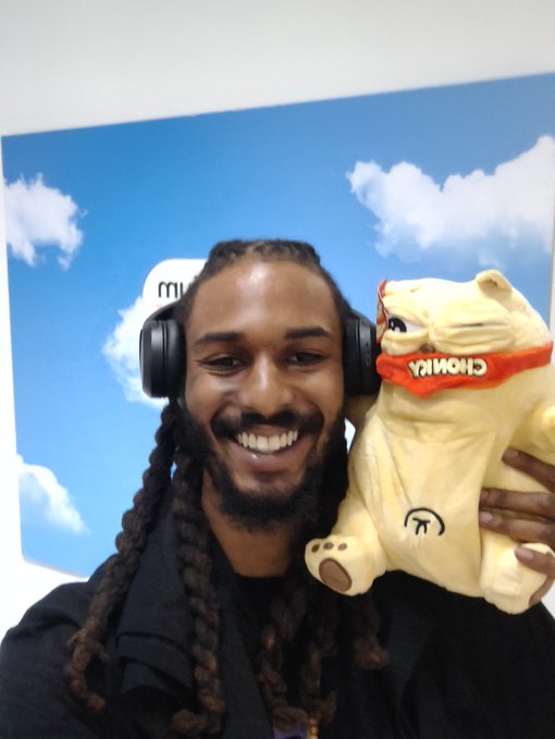 A smiling man with long dreadlocks wearing headphones and a black jacket, holding up a plush toy with a red bandana that reads 'CHONKA'.