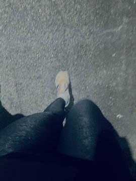 A first-person perspective looking down at someone's feet wearing light-colored sneakers and dark pants while standing on asphalt.