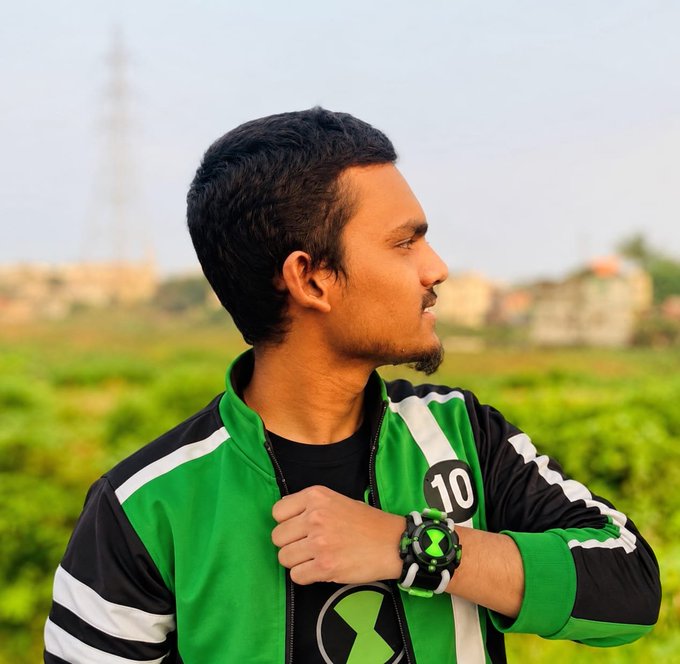 A young man wearing a green and black jacket over a Ben 10 shirt looks to the side, showing off a green Ben 10 themed watch on his wrist against a blurred green background.