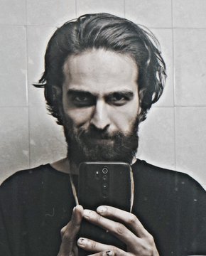 A black and white portrait of a bearded man looking down at a smartphone he is holding in front of his face, taking a mirror selfie.