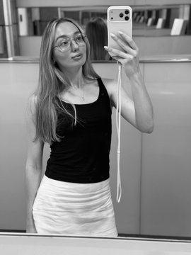 A black-and-white mirror selfie shows a young woman with glasses taking a photo with her smartphone while wearing a black tank top and light skirt.