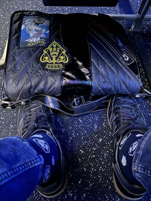 A first-person view looking down at a person's feet wearing black sneakers, with a Disney D23 2025 themed bag featuring a Goose Lounge patch resting on the speckled blue floor between their legs.