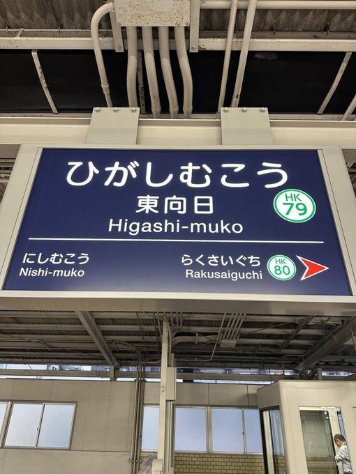 The image shows a sign with Japanese writing, which appears to be located inside an enclosed space or station. The sign has Asian characters written in two different languages, indicating that it might be in a foreign country. A person can also be seen in the background of the image, possibly waiting for transportation or exploring the area around the station.