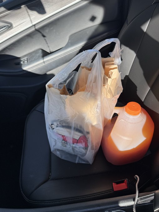 A white plastic bag of food items and an orange drink bottle sitting on a black leather car seat.
