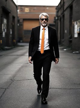 A serious-looking man in a dark suit with a bright orange tie walks down an urban alleyway, his confident stride visually echoing the bullish conviction in the post's claim that $MSTR has bottomed at $82. The stark orange tie against the otherwise desaturated scene symbolizes a flash of optimism amid the bearish context of the post.