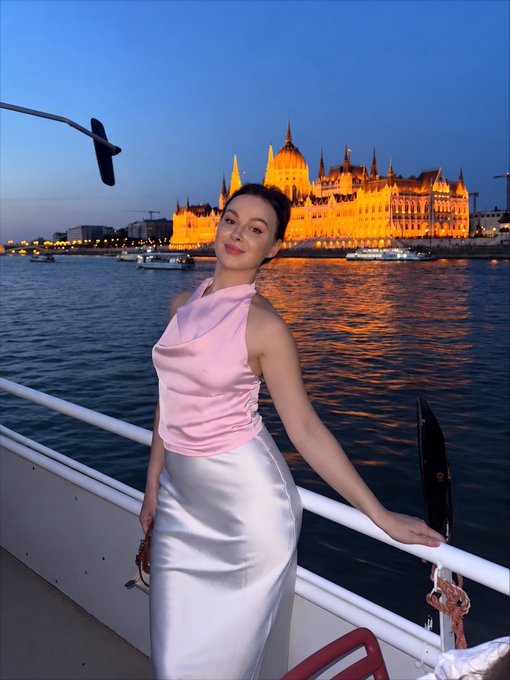 A woman in a pink halter top and white skirt stands on a boat deck at dusk, with the illuminated Hungarian Parliament Building visible across the river in Budapest.