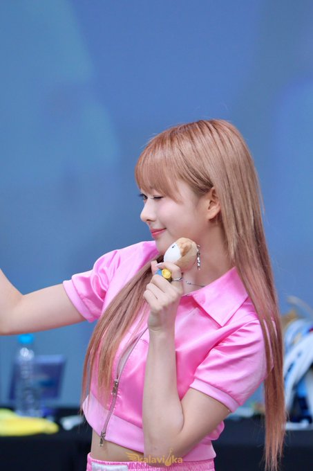 A K-pop idol with long strawberry-blonde hair wearing a pink crop top, smiling while holding a small plush toy during what appears to be a fan event.