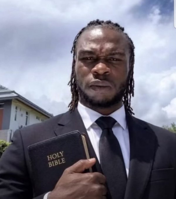 A man in a black suit and tie holding a 'HOLY BIBLE' against his chest, standing outdoors with a cloudy sky and a building visible in the background.