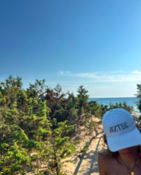 A casual outdoor selfie of a woman wearing a white 'RUTTE' cap on a sunny beach with the ocean and sky in the background.