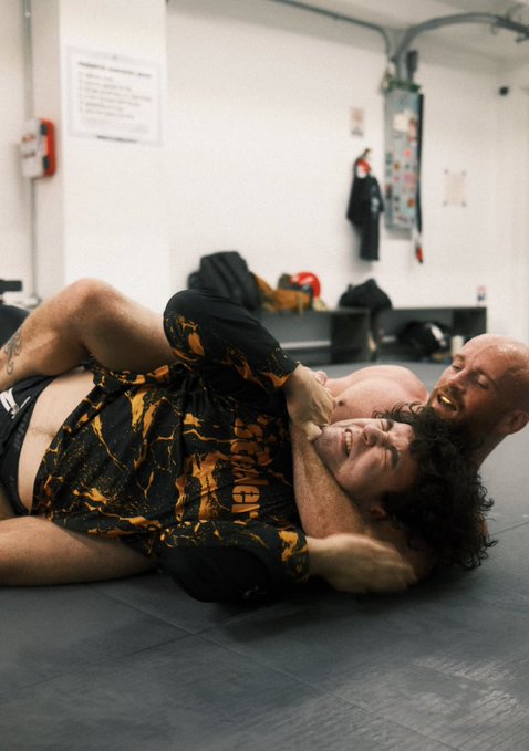 Two men in athletic wear grapple on mats in what appears to be a training gym setting.