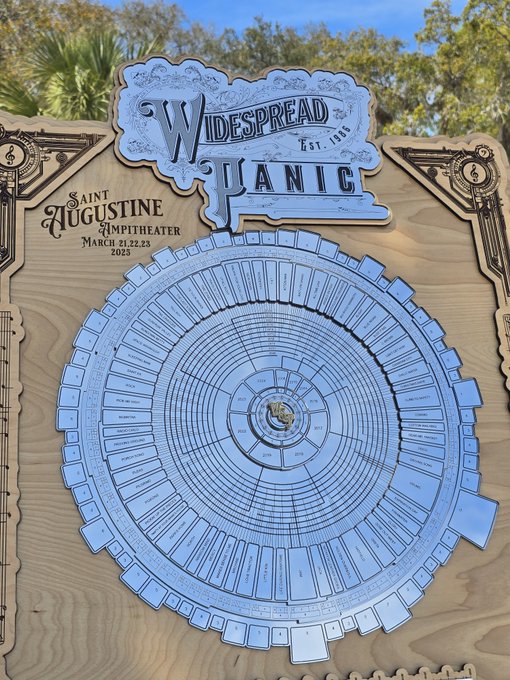 A wooden monument for 'WIDESPREAD PANIC EST. 1986' at 'SAINT AUGUSTINE AMPITHEATER, MARCH 21,22,23 2023' featuring a circular metal disc listing songs and setlist dates.
