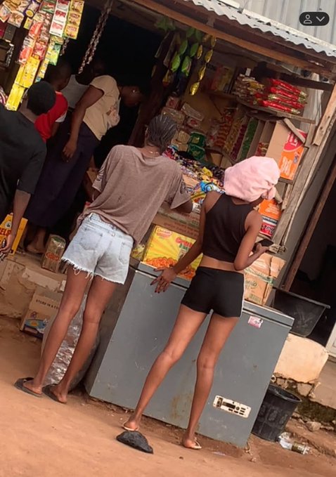 Two young women in shorts and a pink headscarf lean over a gray chest freezer outside a small roadside shop stocked with snacks and Coca-Cola crates, while other customers browse the goods on display.

The caption plays on the post's slang—"See una idols" ("look at you all, idols")—mocking the women's revealing outfits as if they were posing for attention in an everyday neighborhood setting.