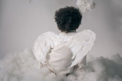 An artist kneels in a field of white feathers or cotton-like clouds, seen from behind wearing a white puffy-sleeved dress.