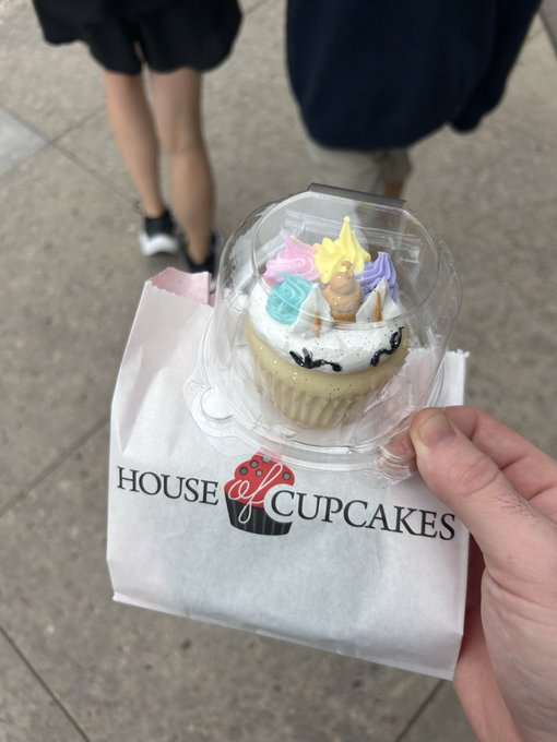 A hand holds a decorated cupcake in a clear plastic container along with a white napkin featuring the "House of Cupcakes" logo. The pastel-frosted cupcake with sprinkles ties in with the "Happy Weekend GM" post as a sweet treat to enjoy over the weekend.