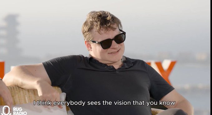 A man with curly hair and sunglasses sitting in a wicker chair during what appears to be a podcast interview, with the subtitle 'I think everybody sees the vision that you know' and a 'RUG RADIO' logo in the corner.
